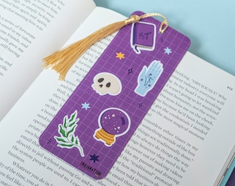 May include: Purple bookmark with a gold tassel. The bookmark features various stickers, including a skull, a crystal ball, a hand with a magic symbol, and a book with the word "Magic" on it. The bookmark also has a green leaf sticker and a star sticker.