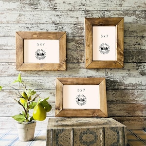 May include: Three rustic, wooden picture frames, each measuring 5 x 7 inches, are displayed against a weathered wood background. The frames have a natural wood finish and feature the text "5 x 7" and a logo. A small potted plant with green leaves and yellow fruit is in the foreground.