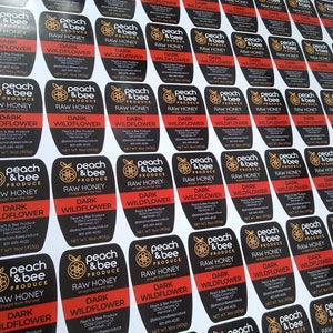 May include: A close-up of multiple black and red labels with the text "Peach & Bee Produce" and "Raw Honey" printed on them. The labels are printed with a bee and peach design and the text "Dark Wildflower" in white. The labels are 1.6 ounces (45 grams).