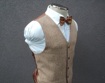 May include: A light brown herringbone vest featuring a V-neckline and four brown buttons down the front. The vest is worn over a light blue collared shirt and paired with a brown patterned bow tie. The garment is displayed on a mannequin torso.