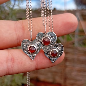 May include: Three silver heart-shaped pendants with a red gemstone centre, each suspended from a delicate chain. The pendants have etched star and crescent moon designs. The jewellery is displayed against a blurred background.