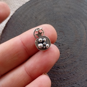 Puede incluir: Un amuleto de plata con forma de granada con una estrella en la parte superior. El amuleto está detallado con pequeñas cuentas redondas que representan las semillas de la fruta.