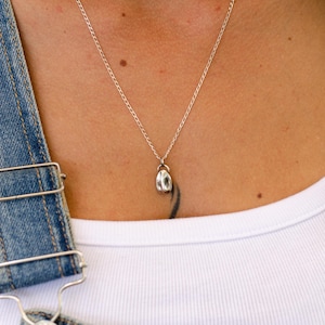 Könnte beinhalten: Eine silberne Halskette mit Anhänger und einem kleinen, abgerundeten Anhänger. Die Halskette wird über einem weißen T-Shirt und einer Jeans-Latzhose getragen. Der Anhänger hat eine strukturierte Oberfläche und hängt an einer feinen Kette.