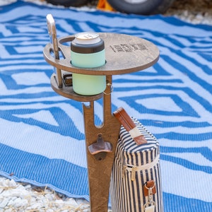 May include: A dark brown wooden stake table with a circular top featuring a beverage holder, a phone slot, and a metal bottle opener. A light mint green insulated can cooler sits in the holder. A blue and white striped fabric bag hangs from the side.