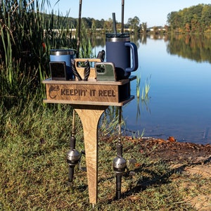 May include: A wooden fishing rod holder with a sign that says "Keepin' It Reel". The holder has two fishing rods attached to it and two blue tumblers. The holder is on the edge of a lake.