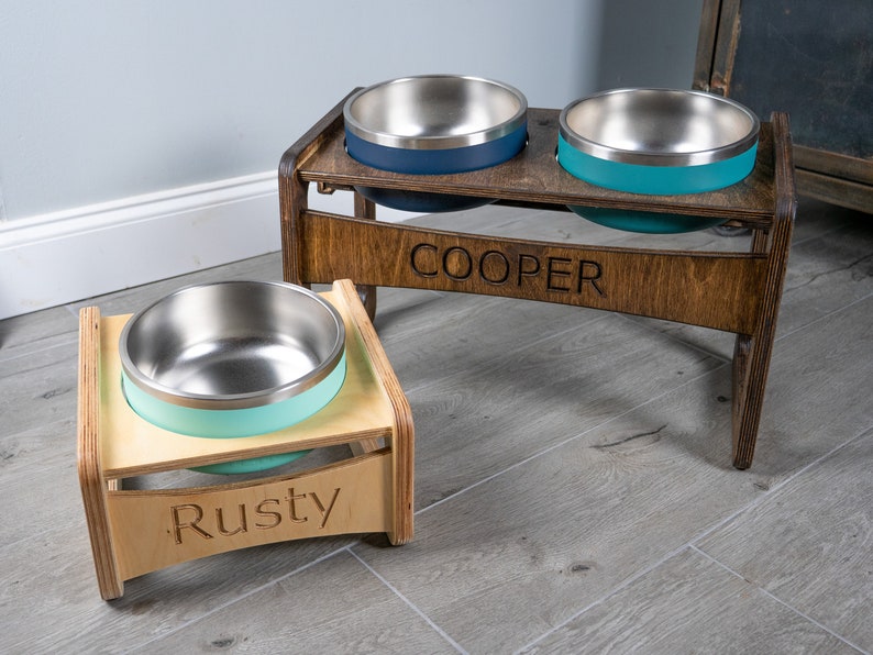 May include: Two elevated dog bowls with stainless steel bowls and a light blue and teal colored exterior. The bowls are on a wooden stand with the names "Rusty" and "Cooper" engraved on them.