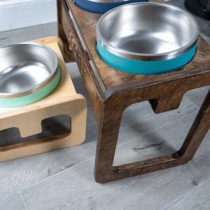 May include: A wooden pet food stand with two stainless steel bowls. The stand has a dark brown stain and the bowls have a light green and teal rim.