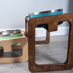 May include: Two elevated pet food bowls, one made of light wood and the other of dark wood. The bowls are stainless steel with a light green rim.