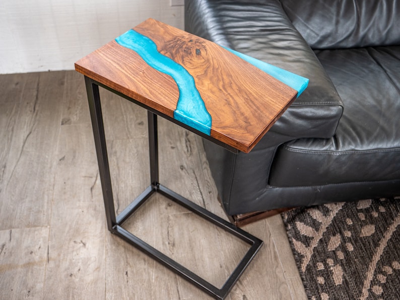 May include: A wooden end table with a blue epoxy river design and a black metal frame. The table is sitting next to a black leather couch.