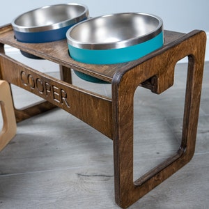 May include: A wooden pet food stand with two stainless steel bowls. The stand is stained a dark brown and has the name "COOPER" engraved on the front. The bowls are teal and blue.