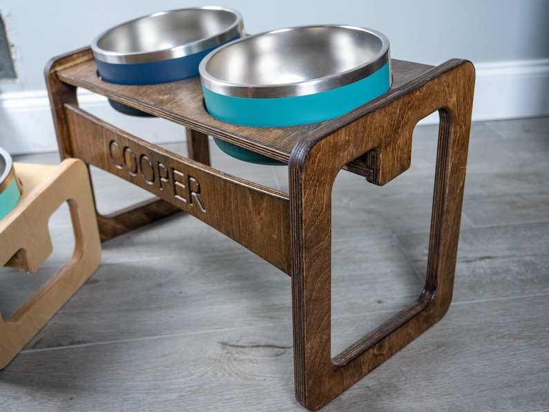 May include: A wooden pet food stand with two stainless steel bowls. The stand is stained a dark brown and has the name "COOPER" engraved on the front. The bowls are teal and blue.