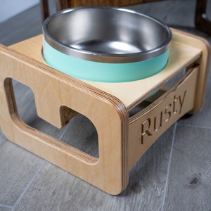 May include: A wooden pet food bowl stand with a light blue bowl and the name "Rusty" engraved on the side.