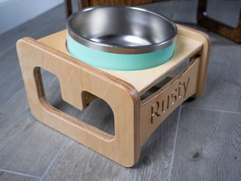 May include: A wooden pet food bowl stand with a light blue bowl and the name "Rusty" engraved on the side.