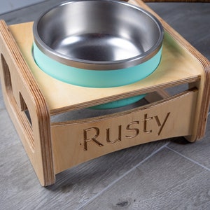 May include: A wooden pet food bowl stand with a light blue stainless steel bowl. The stand is engraved with the name "Rusty".