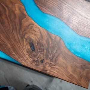 May include: A close-up of a wooden table top with a blue epoxy river running through it. The wood has a natural grain pattern and the epoxy is smooth and shiny.