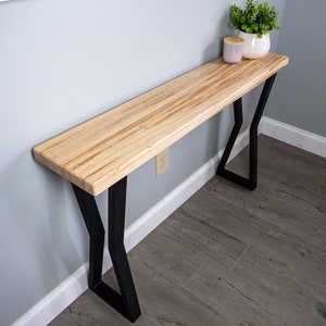 May include: A wooden console table with a natural wood finish and black metal legs. The table has a rectangular top and is supported by two pairs of angled legs.