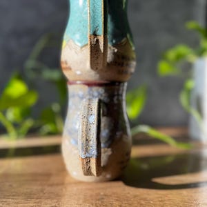 May include: Two stacked ceramic mugs with speckled clay bodies. The top mug features a teal glazed upper section and a tan lower section. The bottom mug has a blue and grey mottled glaze over a tan base. Both mugs have prominent side handles.
