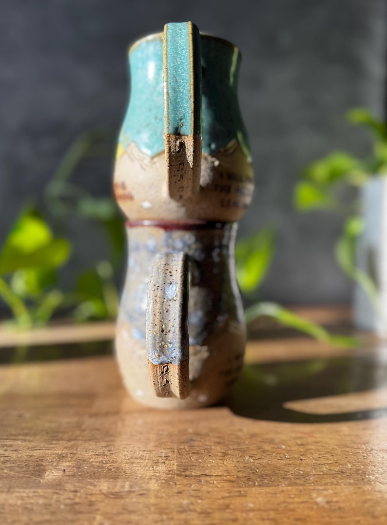 May include: Two stacked ceramic mugs with speckled clay bodies. The top mug features a teal glazed upper section and a tan lower section. The bottom mug has a blue and grey mottled glaze over a tan base. Both mugs have prominent side handles.