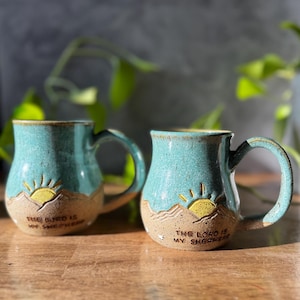 May include: Two ceramic mugs with a speckled teal glaze on the upper half and a textured beige base. Each mug features a carved yellow sun rising over mountains and the embossed text "THE LORD IS MY SHEPHERD" on the lower front section.