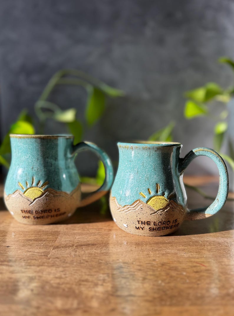 May include: Two ceramic mugs with a speckled teal glaze on the upper half and a textured beige base. Each mug features a carved yellow sun rising over mountains and the embossed text "THE LORD IS MY SHEPHERD" on the lower front section.