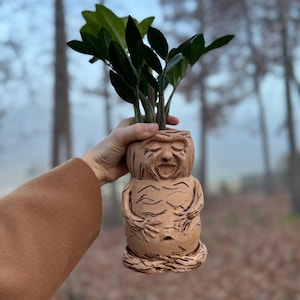 May include: A terracotta planter shaped like a human figure with a face and a textured surface. A green plant is growing out of the top of the planter.