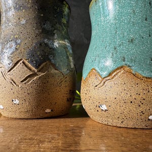 May include: Two ceramic mugs with a speckled tan base featuring carved mountain silhouettes and small painted sheep. The left mug has a dark grey and blue glaze, while the right mug has a bright teal glaze. Both mugs have a textured, earthy finish.
