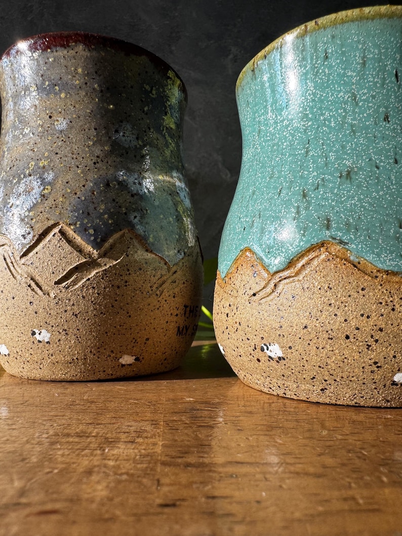 May include: Two ceramic mugs with a speckled tan base featuring carved mountain silhouettes and small painted sheep. The left mug has a dark grey and blue glaze, while the right mug has a bright teal glaze. Both mugs have a textured, earthy finish.