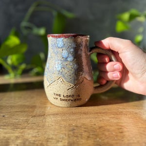 May include: A ceramic mug with a speckled tan base and a blue and white mottled glaze on the upper portion. The mug features a carved mountain range design and the text THE LORD IS MY SHEPHERD embossed in black lettering near the bottom base.