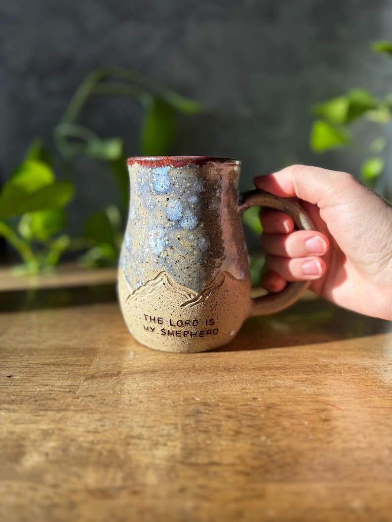 May include: A ceramic mug with a speckled tan base and a blue and white mottled glaze on the upper portion. The mug features a carved mountain range design and the text THE LORD IS MY SHEPHERD embossed in black lettering near the bottom base.