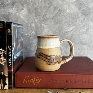 Puede incluir: Una taza de cerámica marrón con borde y asa color crema. La taza presenta un diseño en relieve de una nave espacial. La taza está sobre un libro granate con la palabra "Firefly" en letras doradas.