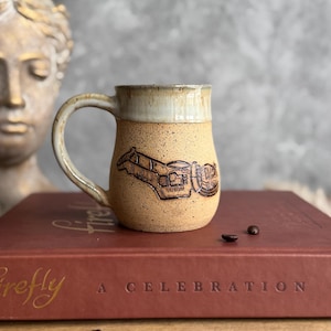 Puede incluir: Una taza de cerámica beige con un diseño grabado detallado. La taza tiene un borde y un asa de color claro. La taza está sobre un libro rojo con las palabras "A Celebration". Granos de café esparcidos.