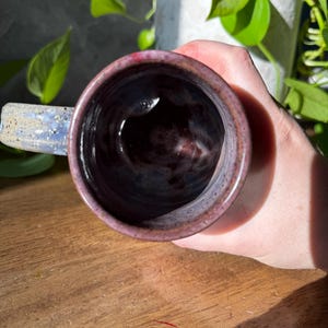 May include: A ceramic mug with a speckled, mottled glaze in shades of deep purple, blue, and dark red. The interior of the mug is dark and glossy, while the exterior features a textured, rustic finish with a sturdy handle extending from the side.