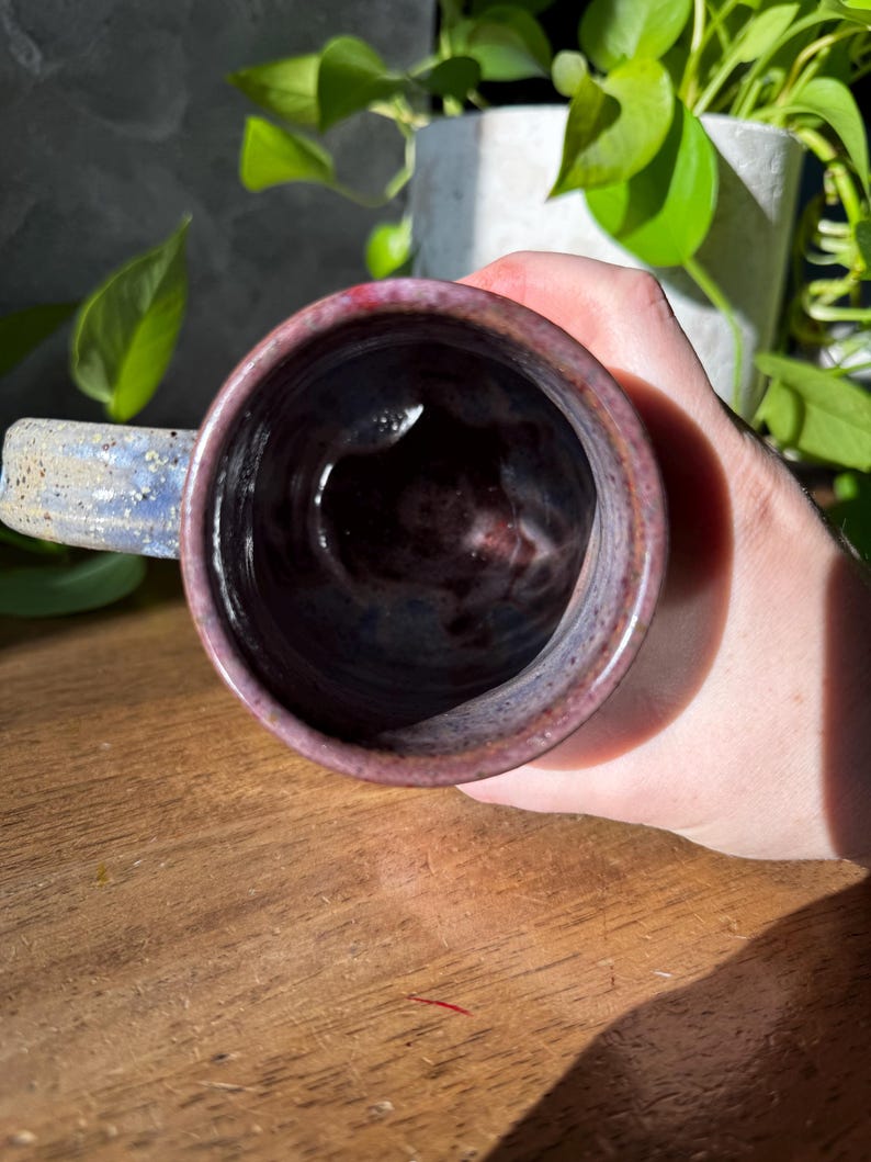 May include: A ceramic mug with a speckled, mottled glaze in shades of deep purple, blue, and dark red. The interior of the mug is dark and glossy, while the exterior features a textured, rustic finish with a sturdy handle extending from the side.