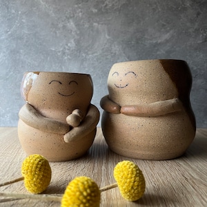 May include: Two ceramic mugs with speckled brown glaze and painted smiley faces. The mugs have arms and hands that are hugging each other. The mugs are sitting on a wooden surface with yellow dried flowers.