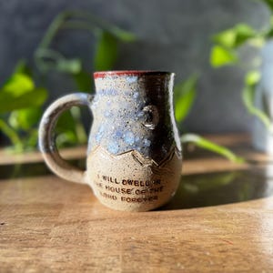 May include: A ceramic mug with a speckled blue and tan glaze, featuring a curved handle and a mountain range relief design. The text "I WILL DWELL IN THE HOUSE OF THE LORD FOREVER" is embossed on the lower front, with a small crescent moon above.