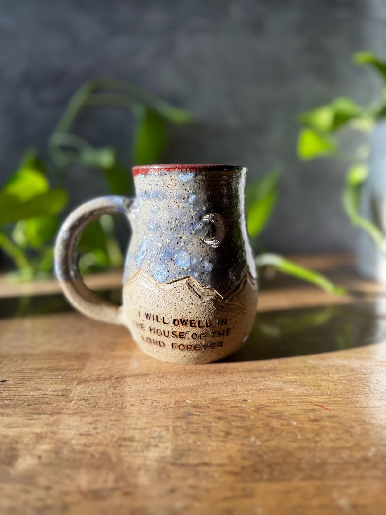 May include: A ceramic mug with a speckled blue and tan glaze, featuring a curved handle and a mountain range relief design. The text "I WILL DWELL IN THE HOUSE OF THE LORD FOREVER" is embossed on the lower front, with a small crescent moon above.