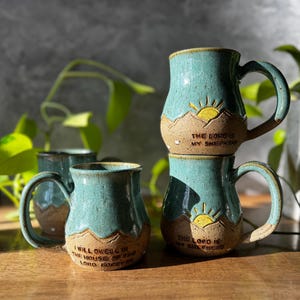 May include: Three ceramic mugs with a speckled teal and tan glaze. Each mug features a raised mountain landscape design with a yellow sun. Two mugs are inscribed with "THE LORD IS MY SHEPHERD" and one with "I WILL DWELL IN THE HOUSE OF THE LORD FOREVER."