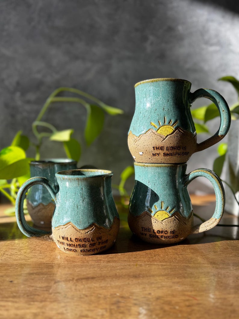 May include: Three ceramic mugs with a speckled teal and tan glaze. Each mug features a raised mountain landscape design with a yellow sun. Two mugs are inscribed with "THE LORD IS MY SHEPHERD" and one with "I WILL DWELL IN THE HOUSE OF THE LORD FOREVER."
