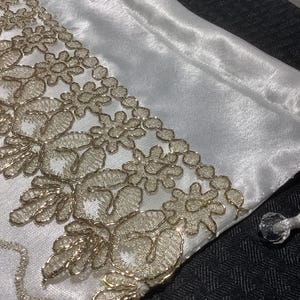 May include: White satin pouch with a gold lace trim. The pouch has a drawstring closure with a crystal bead.