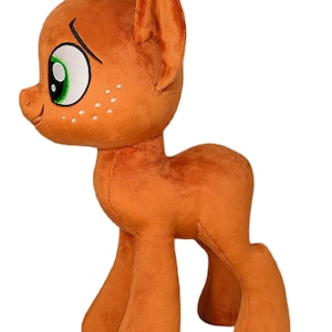 Peut inclure: Un jouet en peluche de poney orange avec des yeux verts et des taches blanches. Le poney est debout sur ses quatre pattes et a une expression amicale.