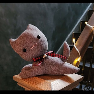 May include: A gray sock monkey cat toy with a red and black plaid bow and a bell. The toy is sitting on a wooden surface.