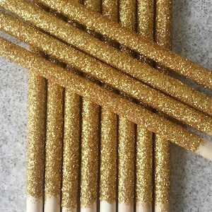May include: A bundle of 12 gold glitter cake pops sticks. The sticks are wooden with a pointed end and are covered in gold glitter.