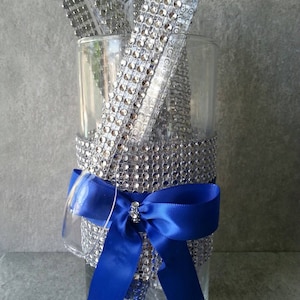May include: A clear glass vase decorated with silver rhinestones and a royal blue satin bow. The vase contains silver rhinestone-covered ribbons. The vase is a decorative item.