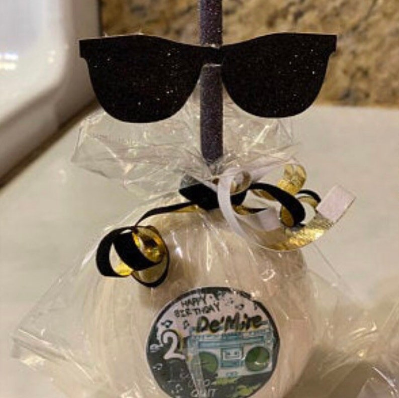 K&ouml;nnte beinhalten: Ein wei&szlig;er Zuckerapfel mit einem schwarz-wei&szlig;en Aufkleber, auf dem "Happy Birthday Be Thirty De'Mire" steht, und einer schwarzen und wei&szlig;en Glitzer-Sonnenbrillen-Dekoration oben drauf.