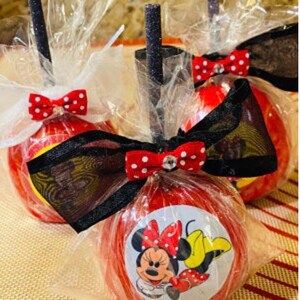 K&ouml;nnte beinhalten: Drei rote Zucker&auml;pfel, verziert mit schwarzen und wei&szlig;en Tupfenb&auml;ndern und schwarzem und wei&szlig;em Band. Ein Apfel hat einen Minnie Mouse-Aufkleber mit einer roten Schleife und einem gelben Kleid.