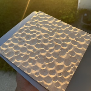 May include: A square wooden panel featuring a textured surface with numerous carved circular indentations. The light-colored wood displays natural grain patterns, and the concave depressions create a repeating, rhythmic pattern across the entire face of the board.