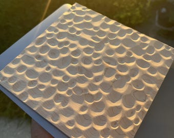 May include: A square wooden panel featuring a textured surface with numerous carved circular indentations. The light-colored wood displays natural grain patterns, and the concave depressions create a repeating, rhythmic pattern across the entire face of the board.