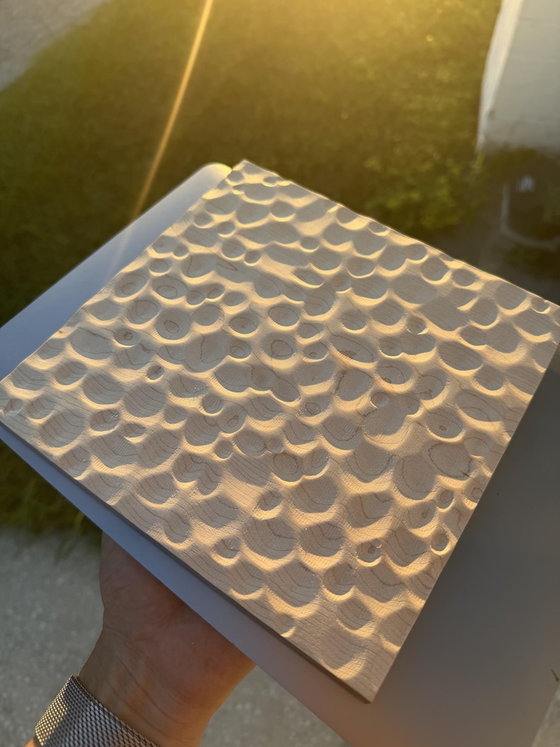 May include: A square wooden panel featuring a textured surface with numerous carved circular indentations. The light-colored wood displays natural grain patterns, and the concave depressions create a repeating, rhythmic pattern across the entire face of the board.