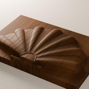 May include: A rectangular wooden block featuring a carved, raised relief of a fan-shaped seashell. The wood displays a rich, medium-brown tone with visible grain patterns running horizontally across the surface and the shell design.