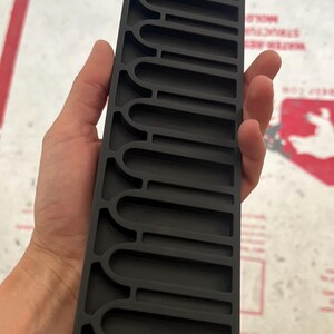 May include: A rectangular black plastic organizer tray featuring a series of uniform, elongated U-shaped slots arranged in a single column. The tray has a matte finish and a structured, rigid design with raised dividers between each individual compartment.